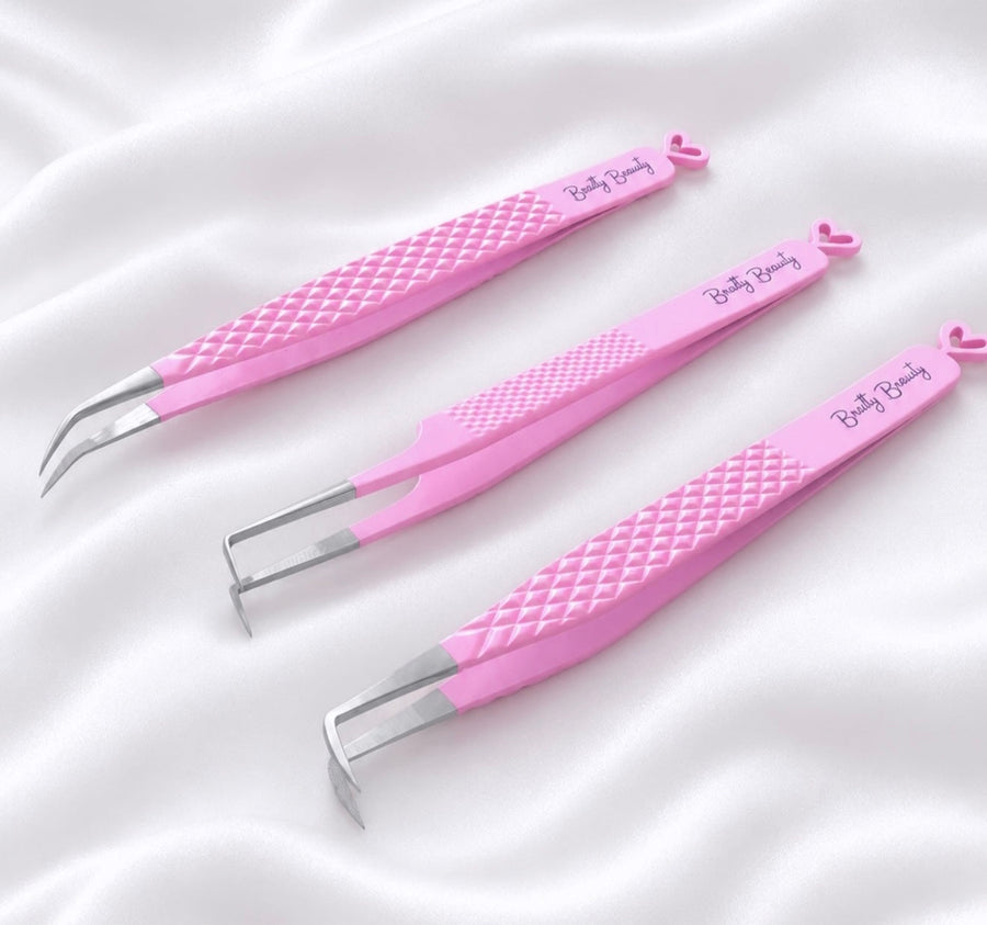 Three pink beauty tweezers with textured grips and silver metal tips, displayed inside a clear acrylic case on a white faux fur background. The tweezers are branded with 'Brattyy Beauty'.Tweezer Trio Set lash volume classic tweezers isolation tweezer