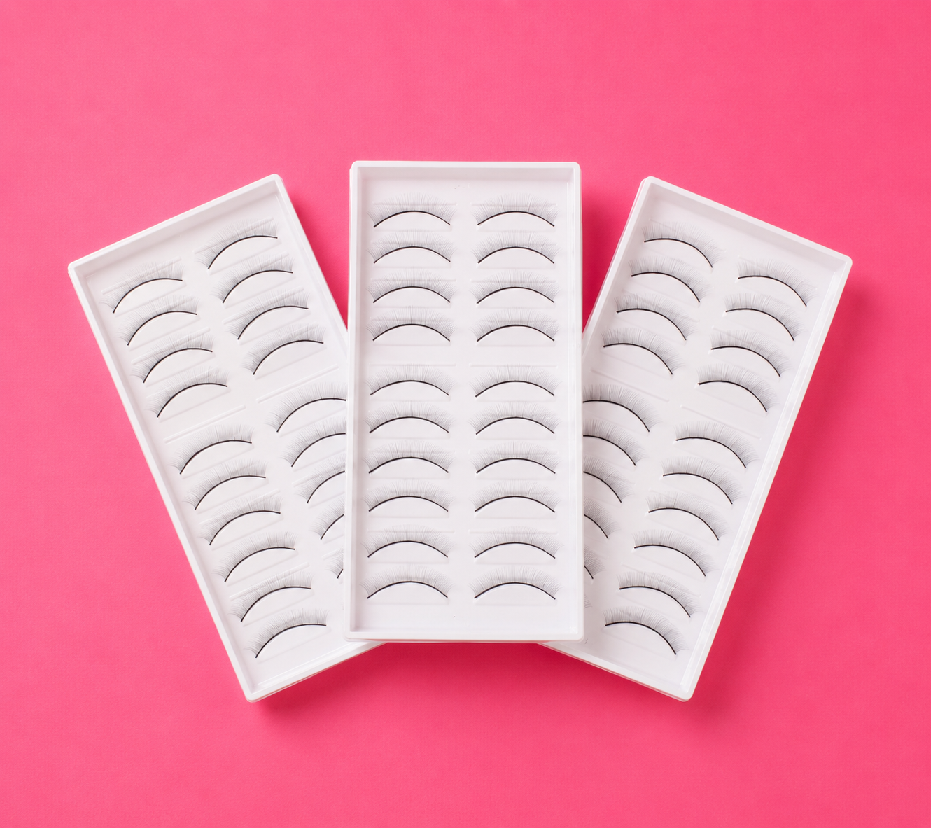 Three white rectangular trays of false eyelashes arranged in a fan shape on a solid pink background. 30 Pairs Of Practice Lash Strips for Mannequin Head | 3 Packs Bratty Beauty XO