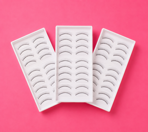 Three white rectangular trays of false eyelashes arranged in a fan shape on a solid pink background. 30 Pairs Of Practice Lash Strips for Mannequin Head | 3 Packs Bratty Beauty XO