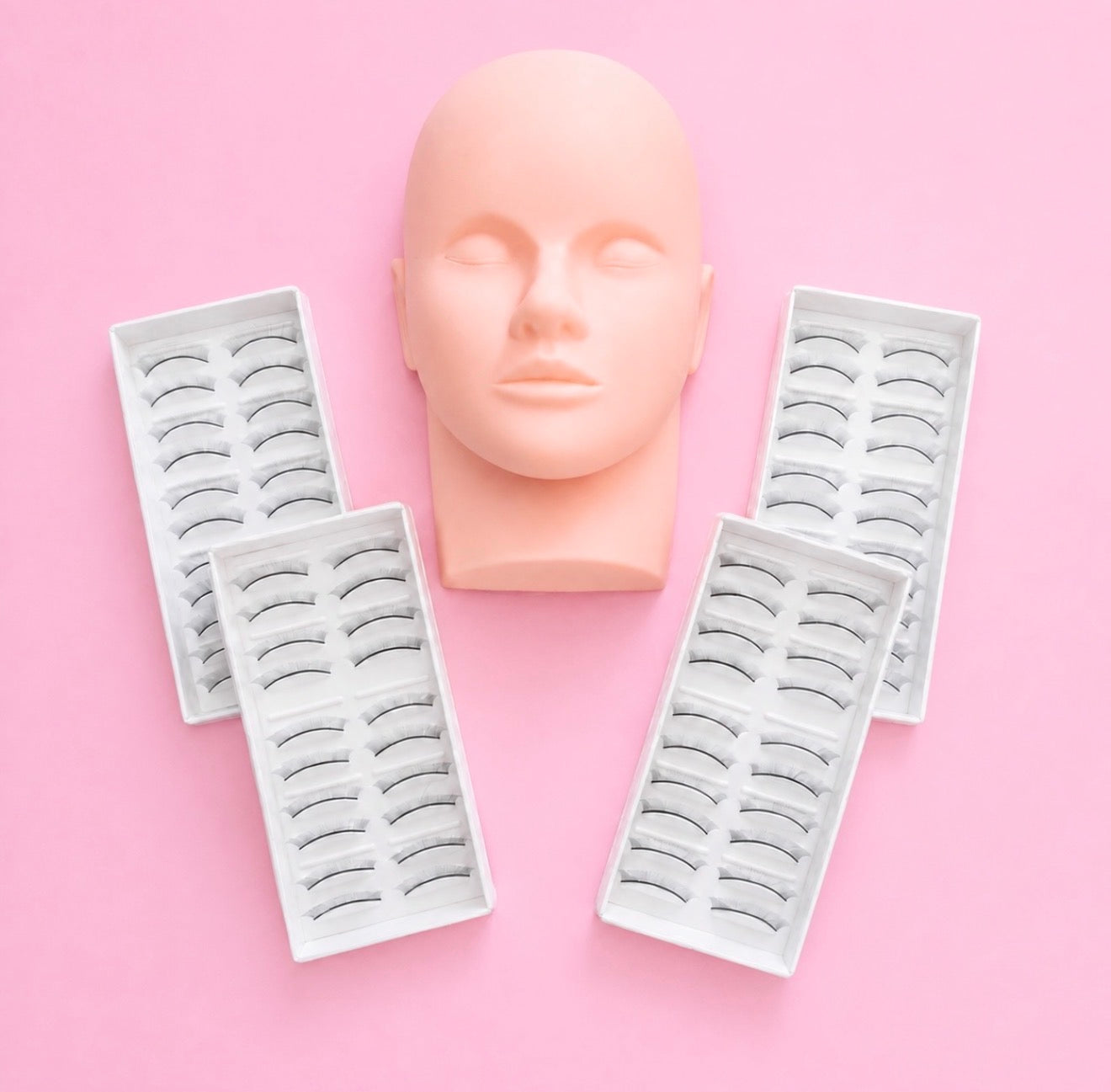 Four white trays of false eyelashes arranged symmetrically around a peach-colored mannequin head on a solid pink background. Lash Artist Practice Starter Pack | A Mannequin Head & 40 Pairs Of Practice Lash Strips Bratty Beauty XO