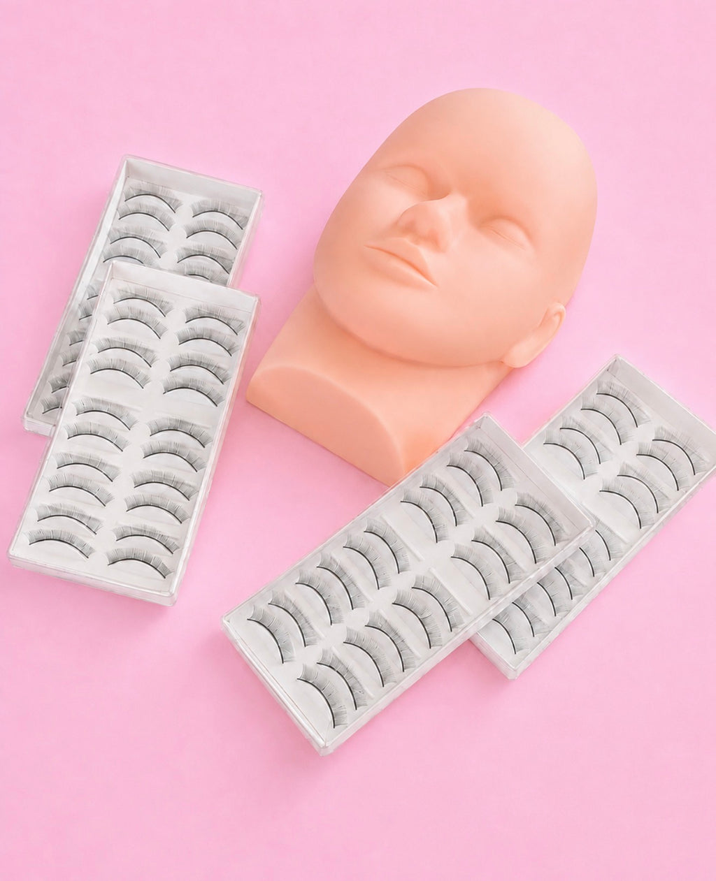 Four white trays of false eyelashes arranged symmetrically around a peach-colored mannequin head on a solid pink background. Lash Artist Practice Starter Pack | A Mannequin Head & 40 Pairs Of Practice Lash Strips Bratty Beauty XO