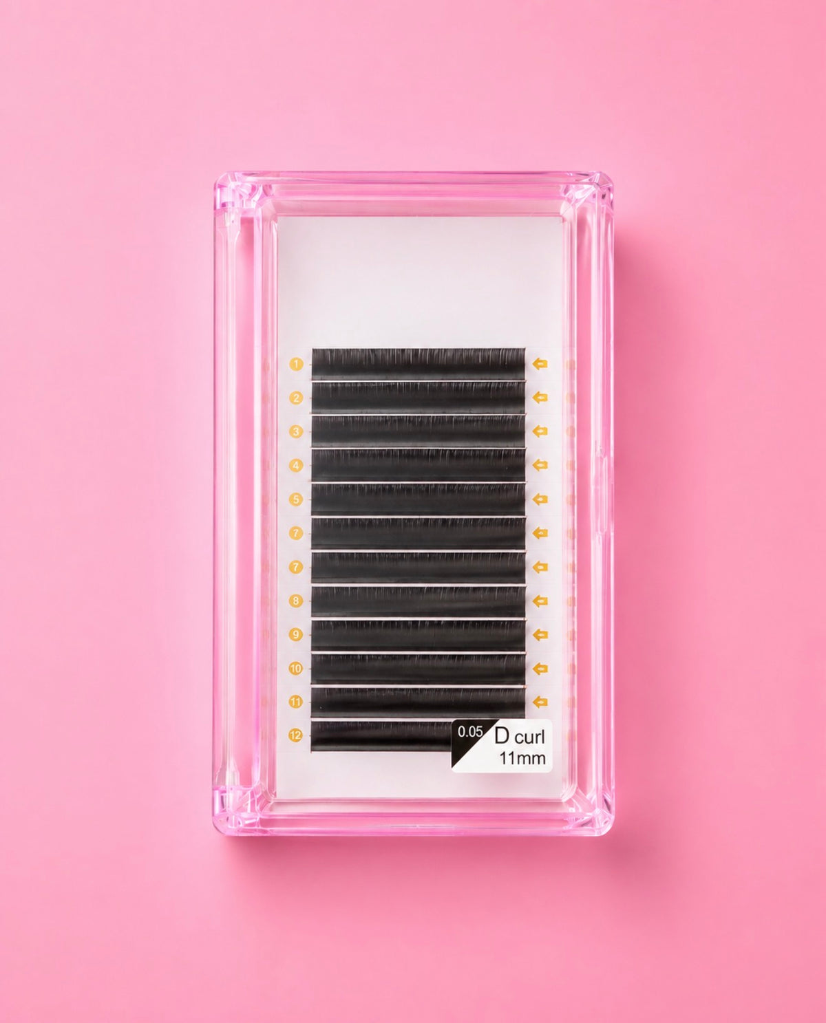 A pack of black false eyelashes in a clear pink plastic case, displayed on a solid pink background. The lash band is labeled 11mm, Matte Black 0.05 D Curl 11mm Lash Extension Tray bratty beauty xo