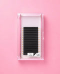 A pack of black eyelash extensions in a clear plastic case, set against a solid pink background. The label on the package reads Matte Black Fast Fanning 0.05 D Curl 13mm Lash Extension Tray bratty beauty xo