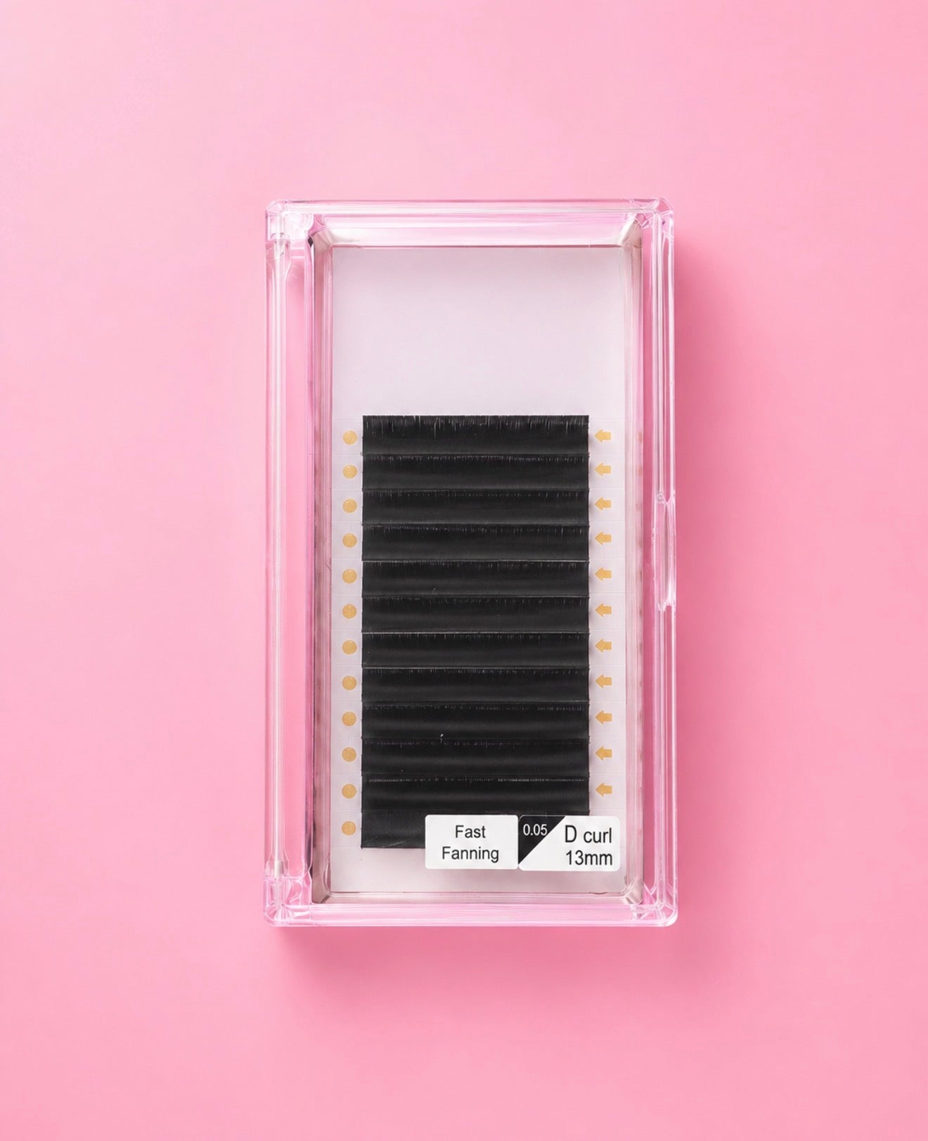 A pack of black eyelash extensions in a clear plastic case, set against a solid pink background. The label on the package reads Matte Black Fast Fanning 0.05 D Curl 13mm Lash Extension Tray bratty beauty xo