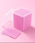 Pink textured square pads inside a clear plastic storage container with one pad removed and fanned out on a solid pink background.Pink or White 400 pcs Lash Adhesive Wipes for Eyelash Extensions