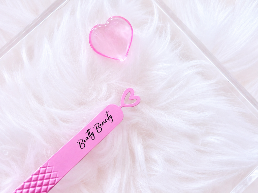 Pink beauty tool set featuring a long spatula labeled 'Butty Beauty' and a small heart-shaped container, displayed on a white faux fur surface.Big Boss Fanning Tweezer, bratty beauty xo volume lash tweezer