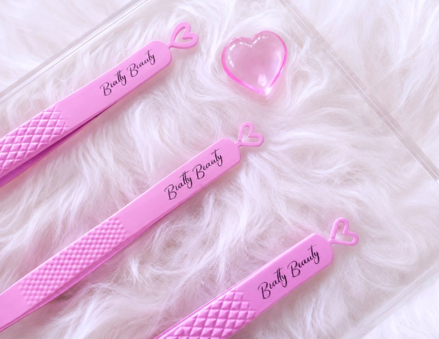 Four pink eyebrow tweezers with textured grips and 'Beauty Beauty' script, arranged on a white faux fur surface next to a small pink heart-shaped accessory.Tweezer Trio Set lash volume classic tweezers isolation tweezer