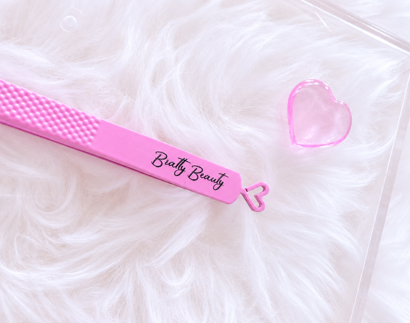 A pink silicone beauty tool with the brand name 'Beatty Beauty' printed on it, resting on a white faux fur surface next to a small clear heart-shaped object.Boss Babe Fanning Tweezer bratty beauty xo volume lash tweezer 