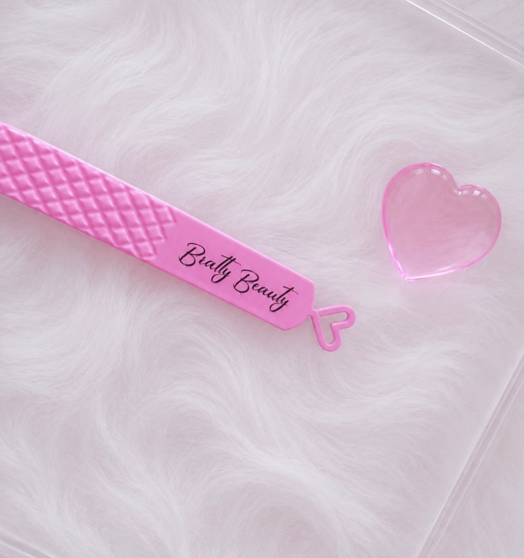 A pink plastic nail file with a diamond-quilted texture and the brand name 'Bratty  Beauty' printed on it, shown next to a small translucent pink heart-shaped piece on a white faux fur background.Boss Isolation Tweezer bratty beauty xo volume lash tweezer