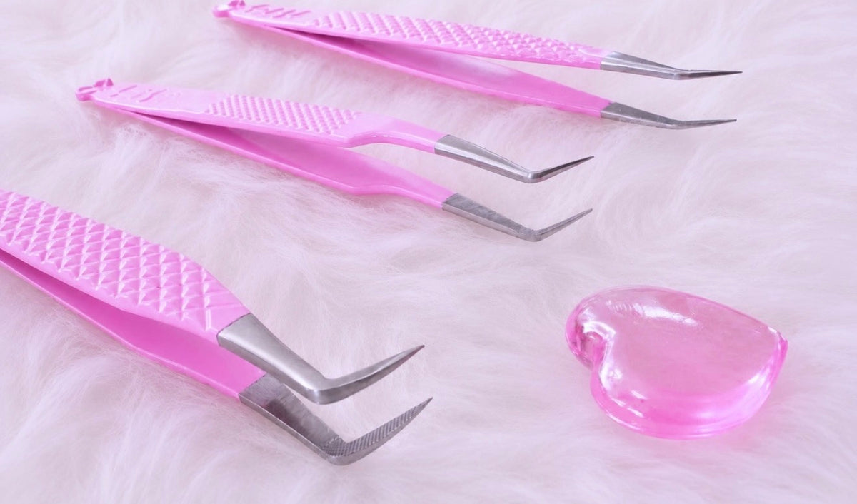 Three pink-handled tweezers with silver metal tips arranged on a white faux fur surface, alongside a small translucent pink heart-shaped object.Tweezer Trio Set lash volume classic tweezers isolation tweezer