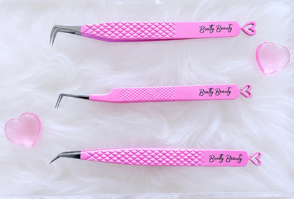 Three pink beauty tweezers with textured grips and silver metal tips, displayed inside a clear acrylic case on a white faux fur background. The tweezers are branded with 'Brattyy Beauty'.Tweezer Trio Set lash volume classic tweezers isolation tweezer