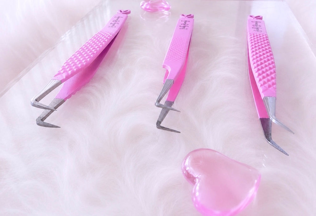 Three pink-handled precision tweezers with silver metal tips arranged on a white faux-fur surface with a small pink heart-shaped object in the foreground.Tweezer Trio Set lash volume classic tweezers isolation tweezer