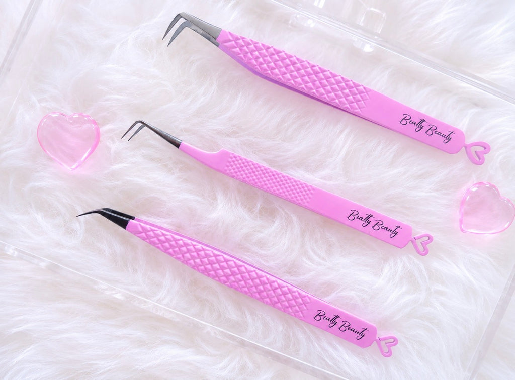Three pink precision tweezers with textured handles and silver metal tips, arranged on a white faux fur surface inside a clear plastic case. The tweezers are branded with 'Body Beauty' and decorated with small pink heart charms.Tweezer Trio Set lash volume classic tweezers isolation tweezer