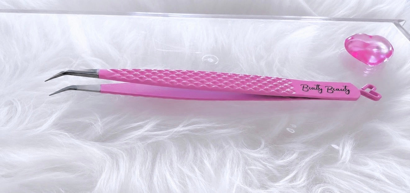 Pink curved tweezers with a textured grip and 'Beauty Beauty' branding, displayed on a white faux fur surface next to a small pink heart-shaped object.Boss Isolation Tweezer bratty beauty xo volume lash tweezer